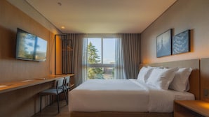 A modern hotel room with a large bed, a desk, and a chair. There is a window with curtains, and two framed pictures on the wall. - Golden Tulip Canela OFICIAL (Canela)