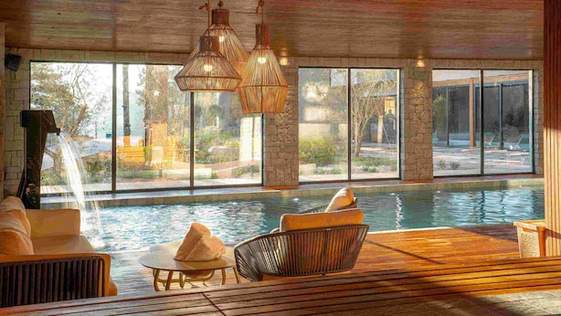 A modern indoor pool area with a waterfall feature, large windows, and comfortable seating. - Golden Tulip Canela OFICIAL (Canela)