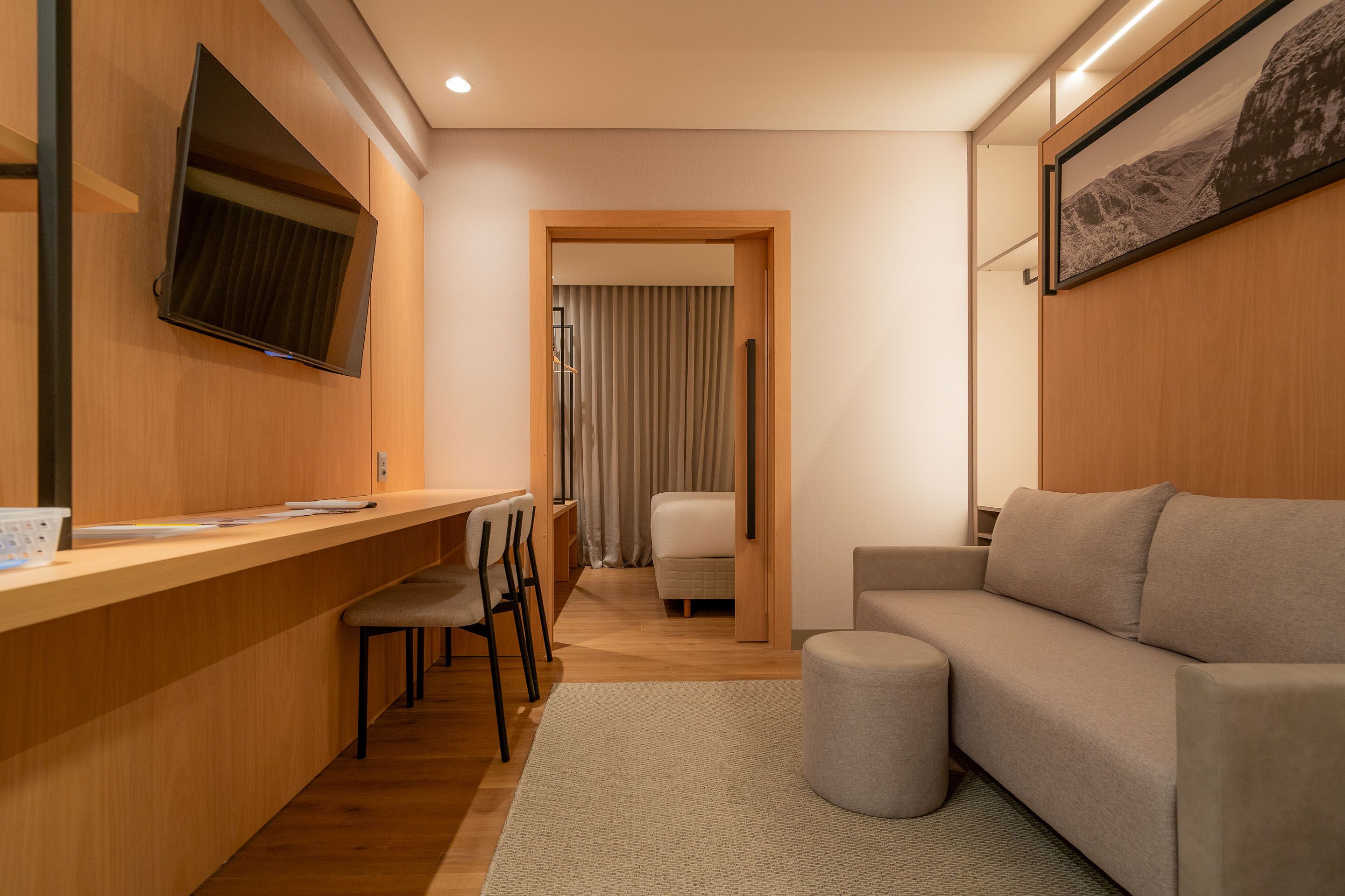 Ein modernes Hotelzimmer mit einem Holztisch, einem flachbildfernseher an der Wand, einer Couch und einem kleinen Hocker.