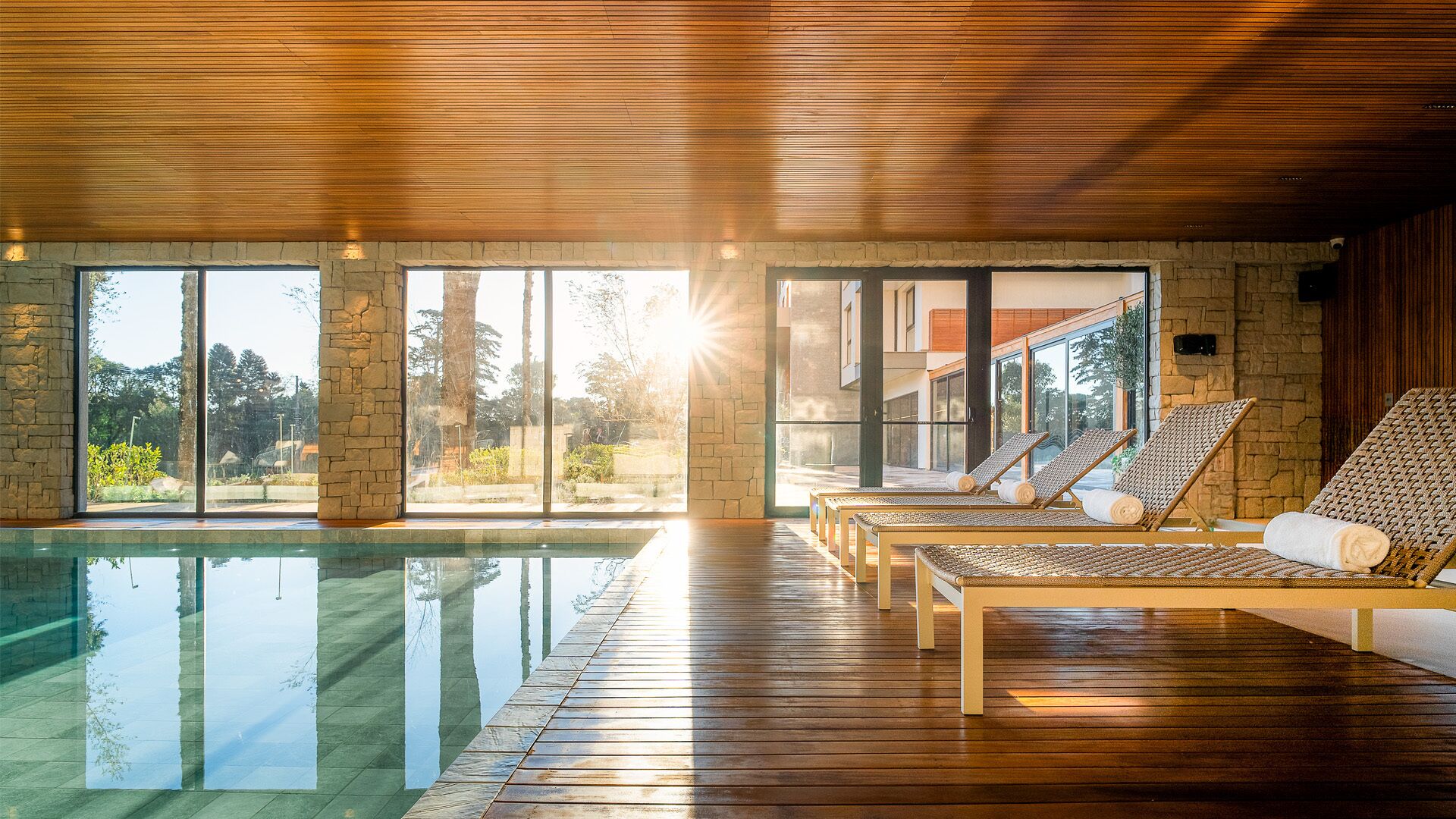 A modern indoor pool area with wooden flooring, stone walls, and large windows offering views of the outdoors.