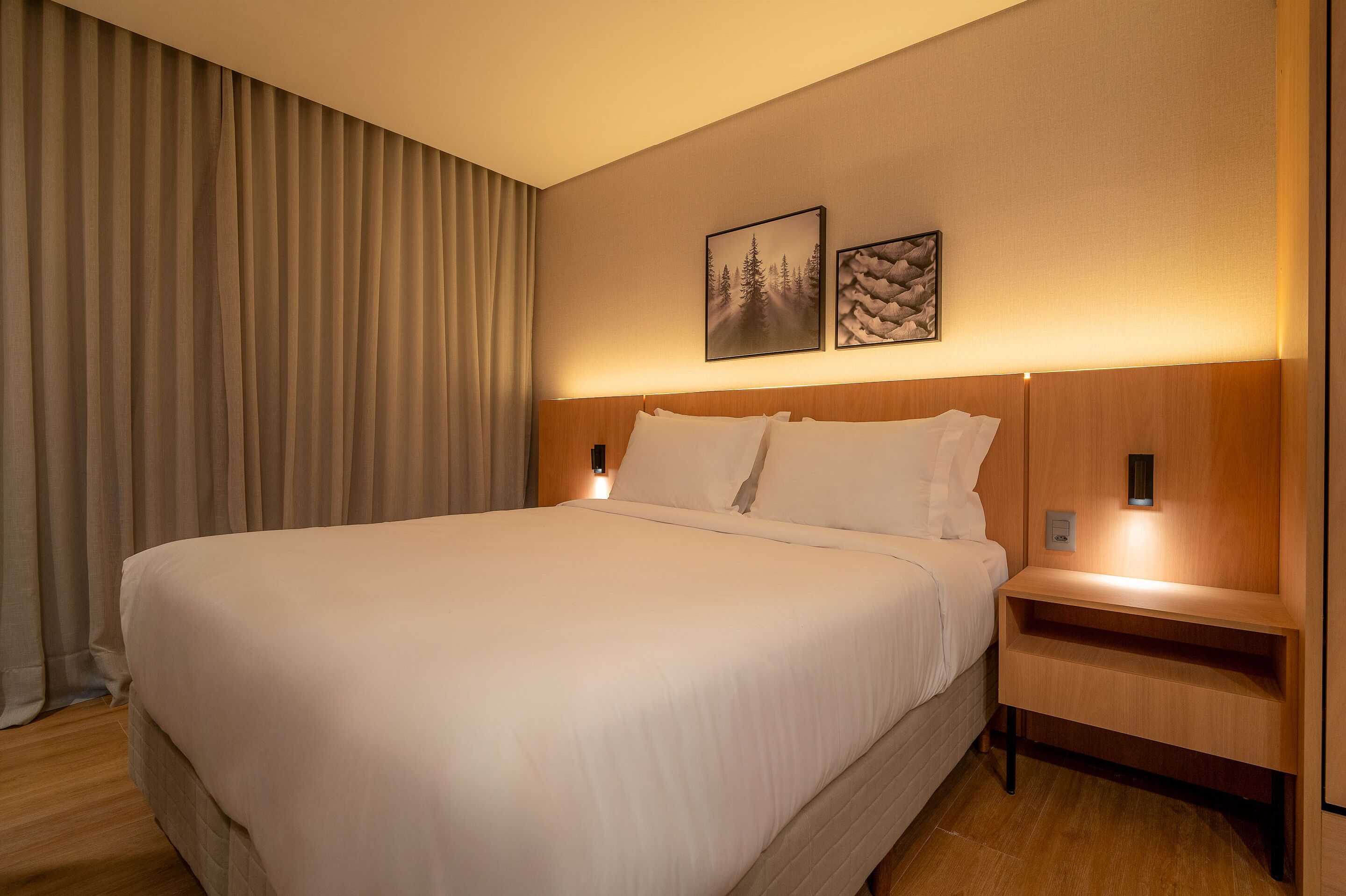 Habitación de hotel con una cama grande, dos cuadros colgados en la pared, una mesita de noche con una lámpara y una mesita auxiliar de madera.