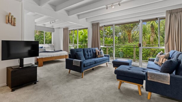 A modern living room with a large flat-screen TV, a sofa, an ottoman, and a bed with a view of lush greenery through large windows.