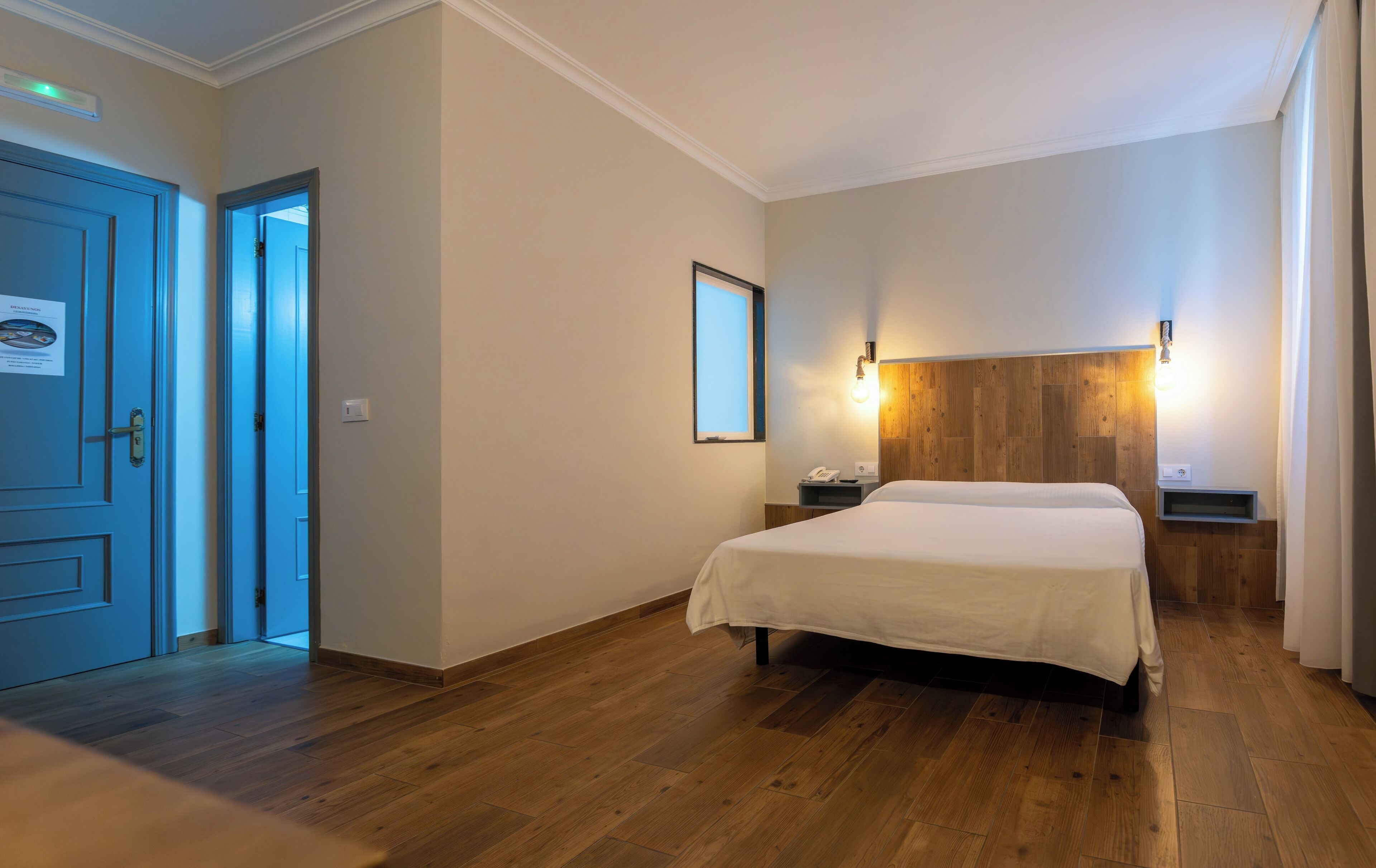 A hotel room with a wooden floor, a bed with a white bedspread, a nightstand with a phone, a wall-mounted light, and a door with a sign.