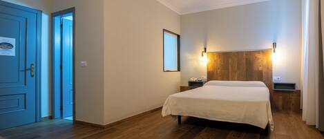 A hotel room with a wooden floor, a bed with a white bedspread, a nightstand with a phone, a wall-mounted light, and a door with a sign.