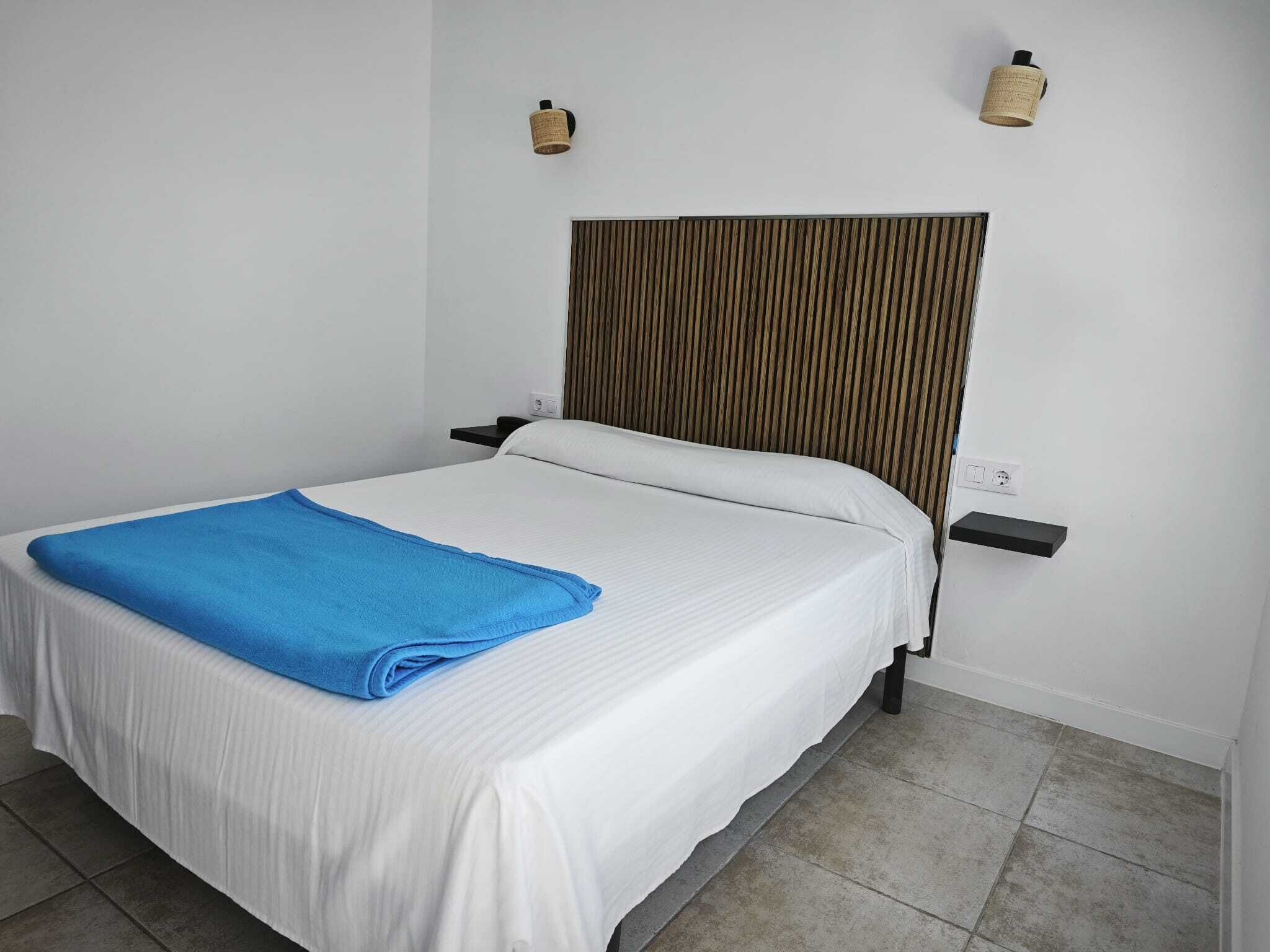 A neatly made bed with a blue towel, two wall-mounted lamps, and a power outlet on the wall.