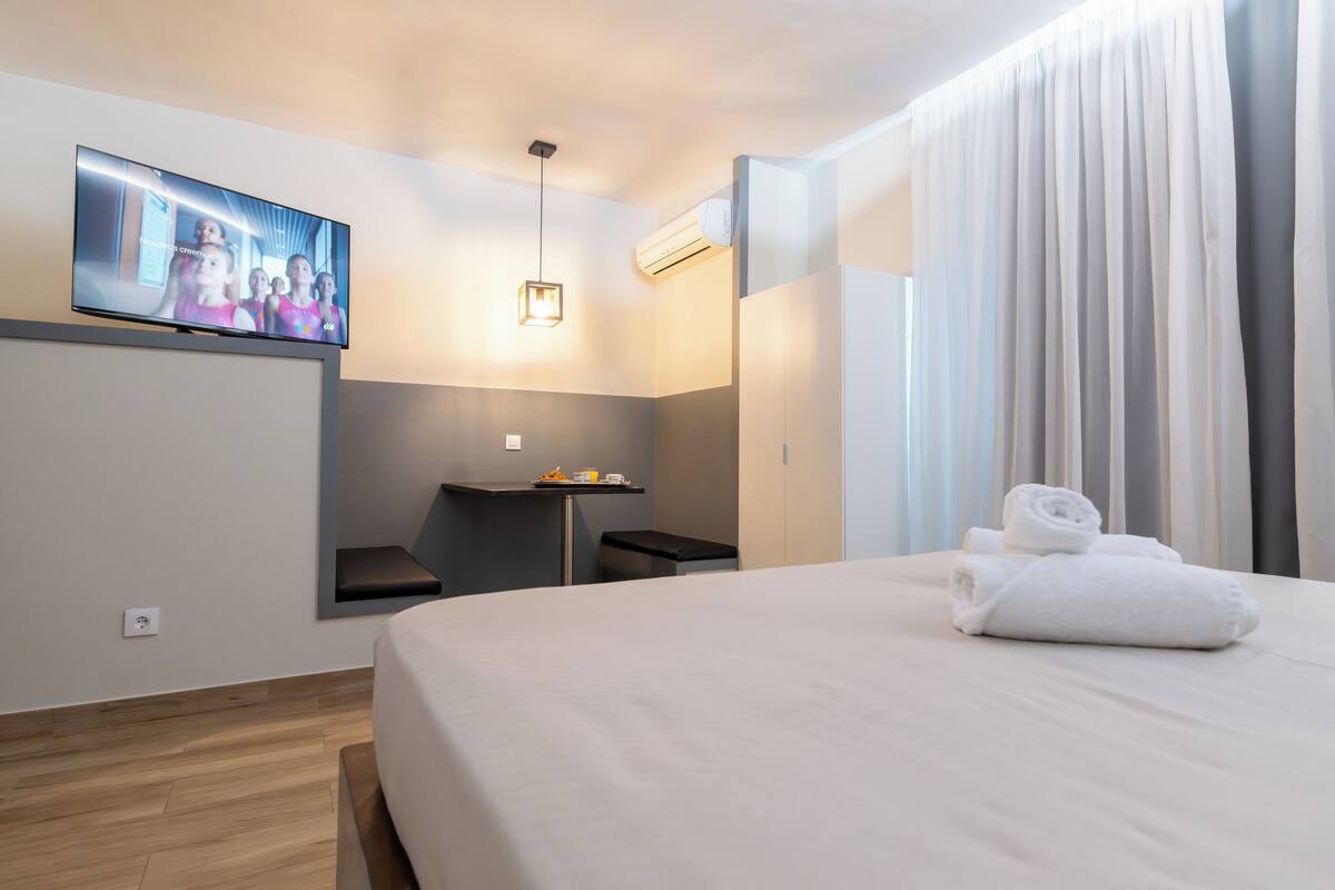 A modern hotel room with a large bed, a flat-screen TV mounted on the wall, a small desk with a chair, and a window with curtains.
