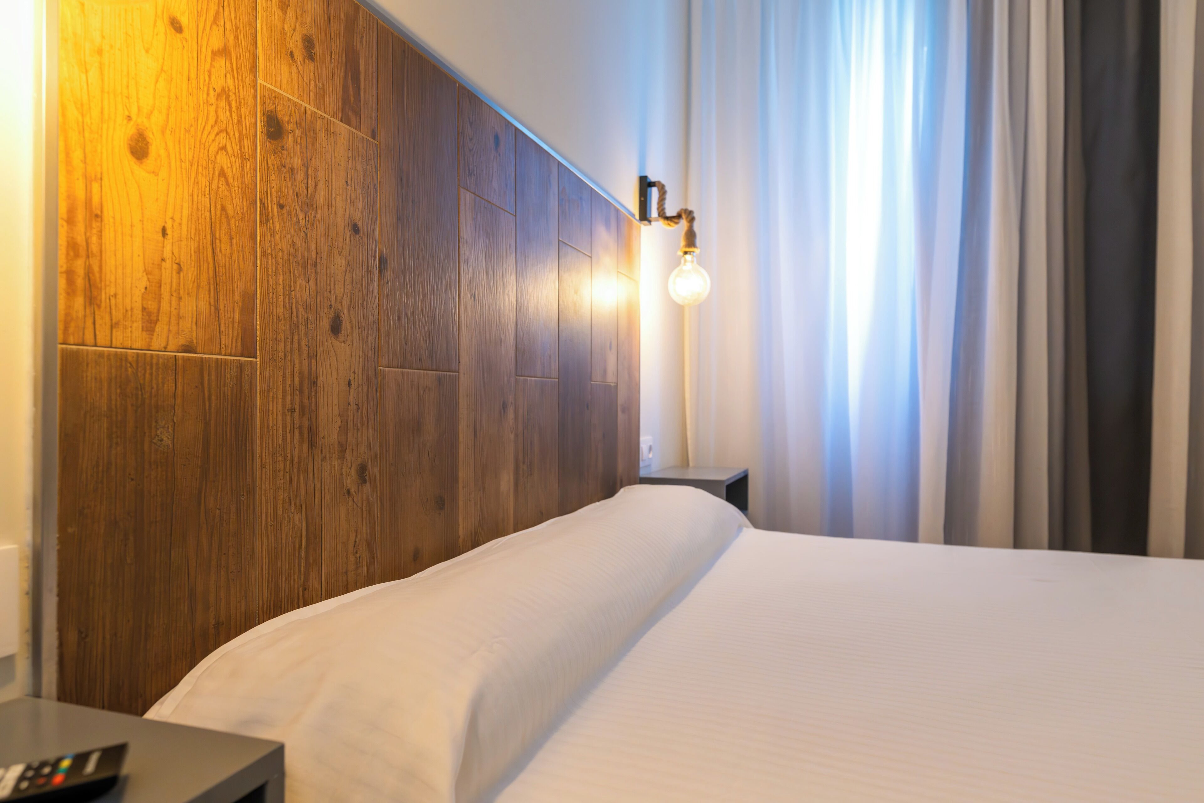 A hotel room with a wooden headboard, a bedside table with a remote control, and a wall-mounted light fixture.