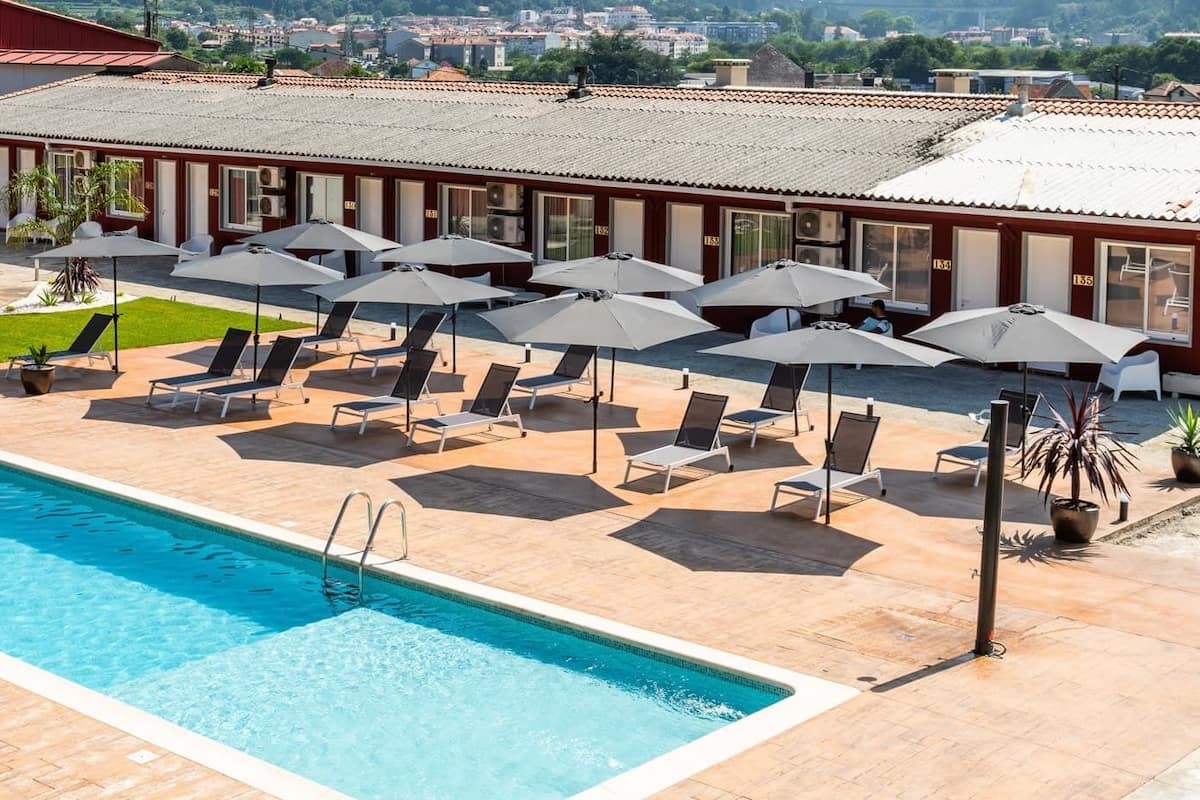 A swimming pool with lounge chairs and umbrellas, surrounded by a red building with multiple windows and a tiled roof.