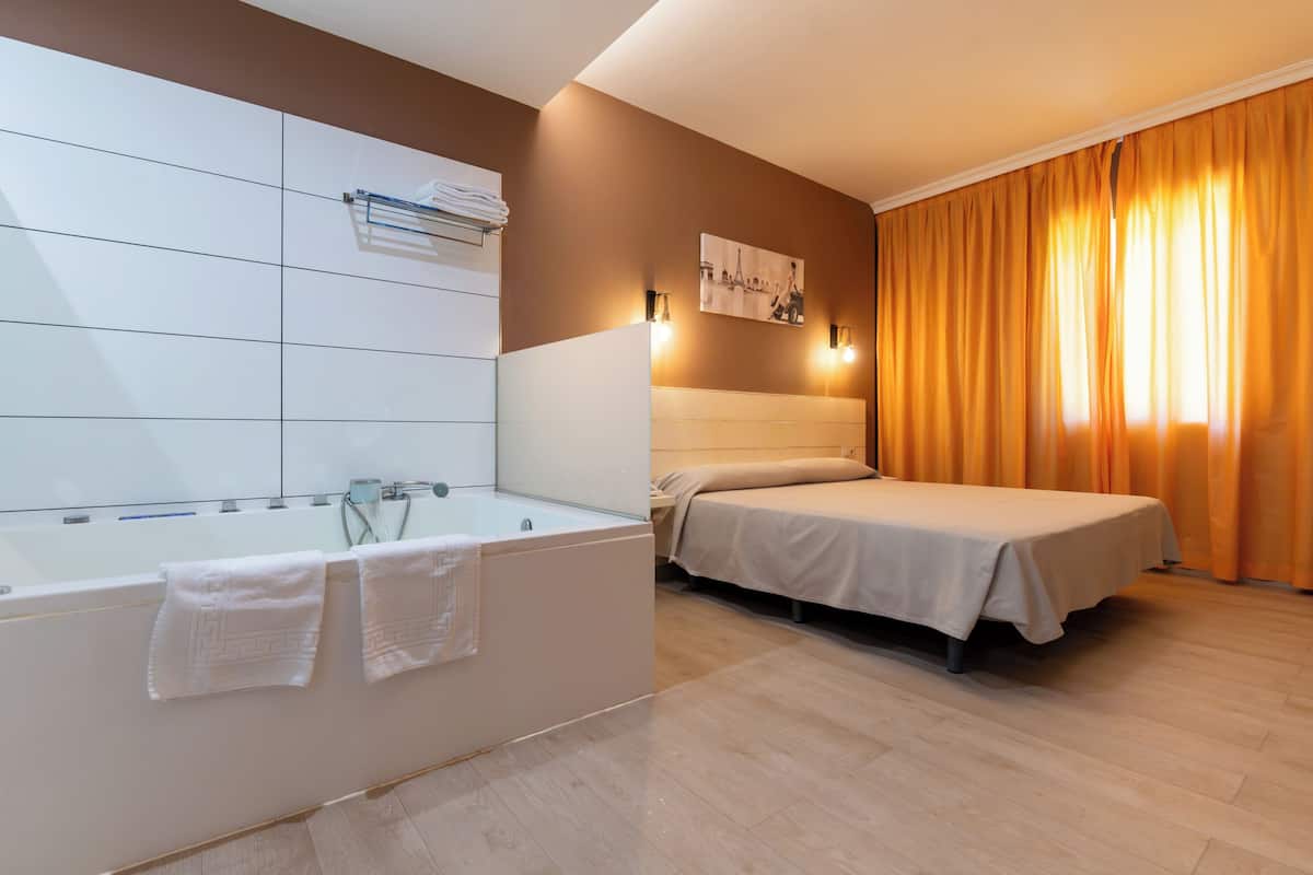 A modern hotel room with a large bathtub, a bed, and a view of the city through curtains.