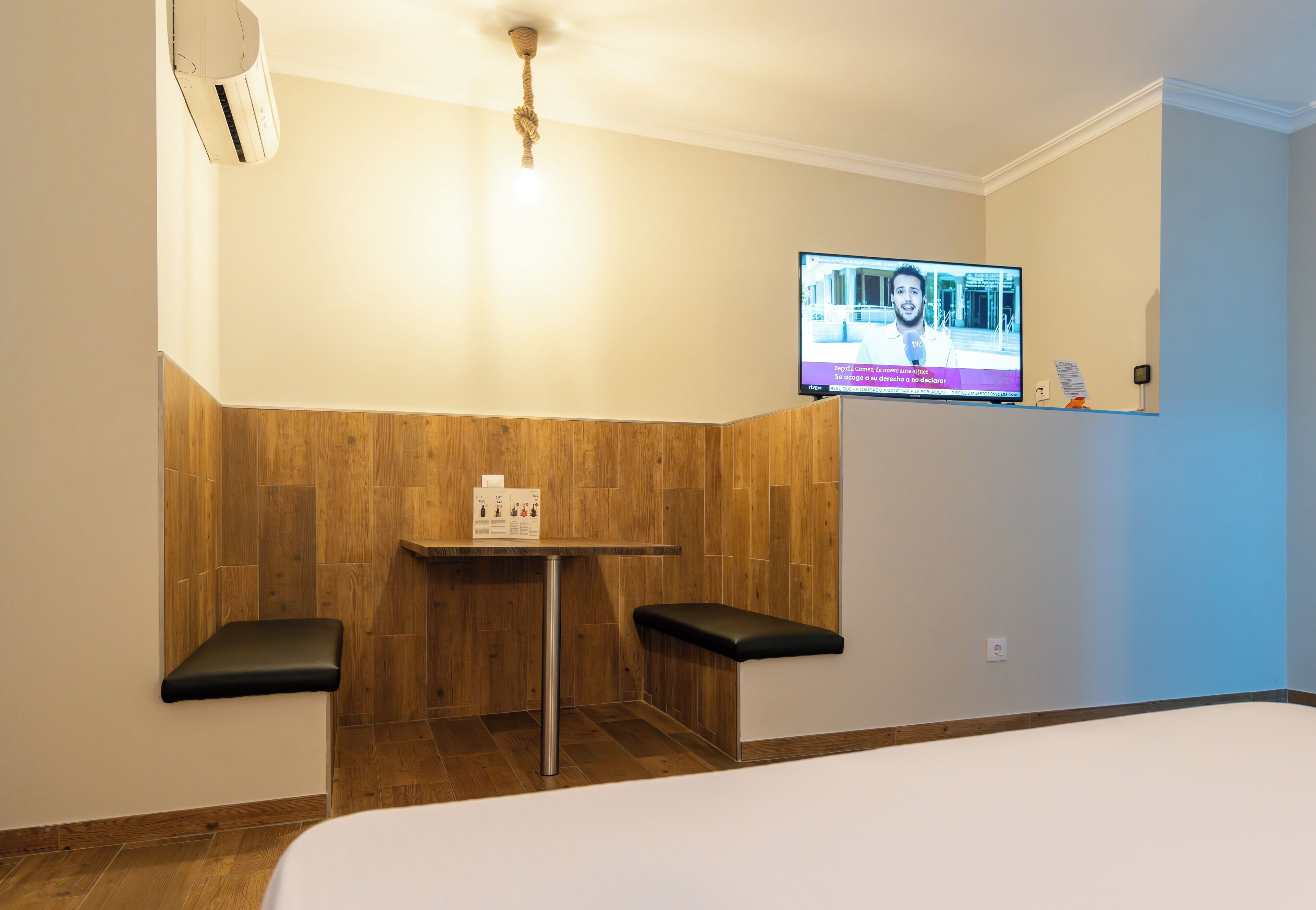 A small, modern room with a wooden wall panel, a single table with a chair, and a television mounted on the wall.
