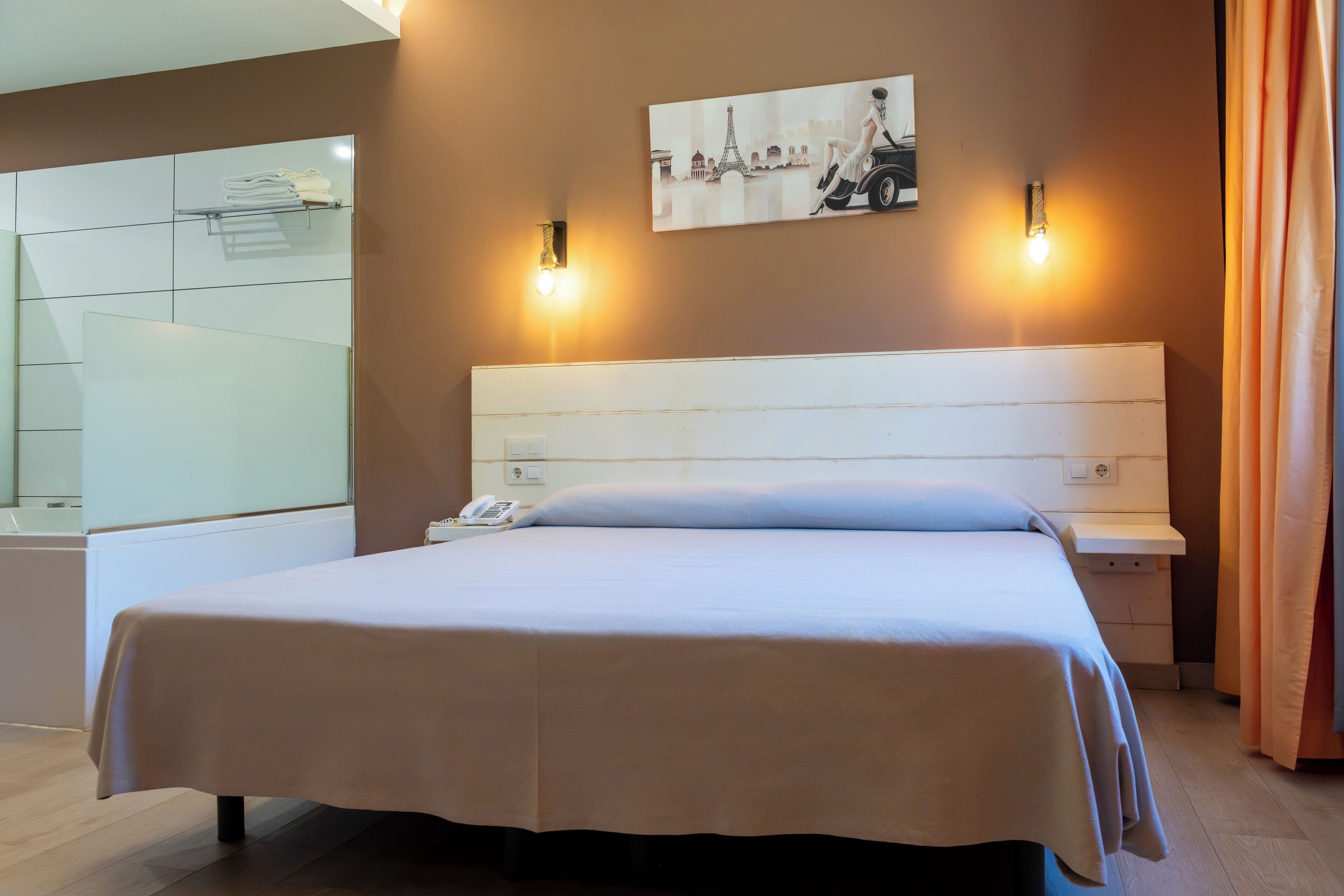 Une chambre d’hôtel avec un grand lit, une table de chevet avec un téléphone, une lampe fixée au mur et un tableau représentant la Tour Eiffel.