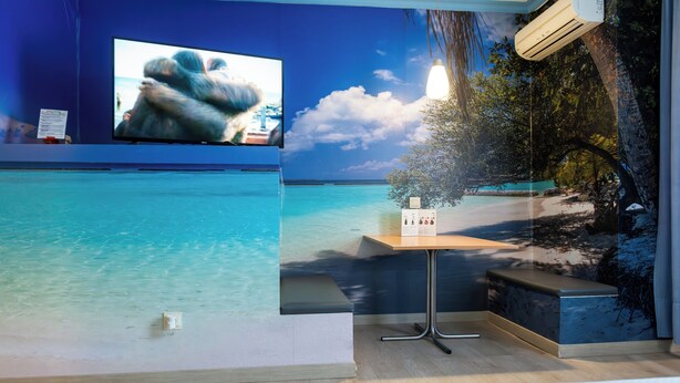 Een kamer met een grote televisie aan de muur, een kleine tafel met een bankje en een strandmuur.