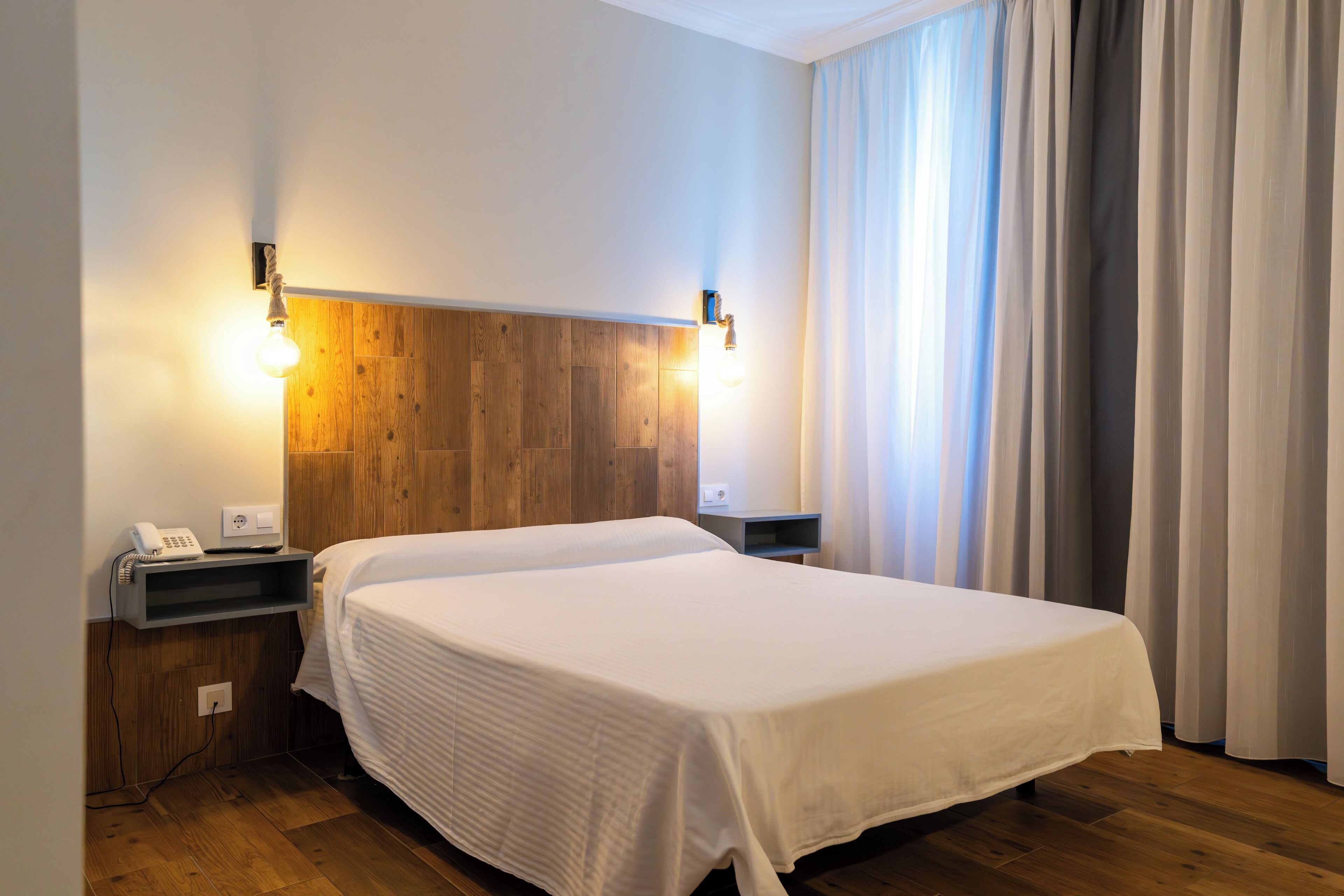 A hotel room with a wooden headboard, a white bedspread, a bedside table with a phone, and a nightstand.