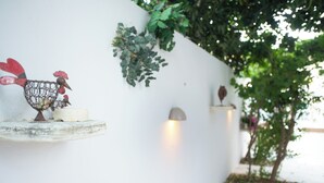 A white wall with a decorative metal rooster and a small bird sculpture on a shelf, a wall-mounted light, and a small tree with green leaves. - Cozy beachside retreat in Kalamaki - steps from the sea (Neo Kalamaki)