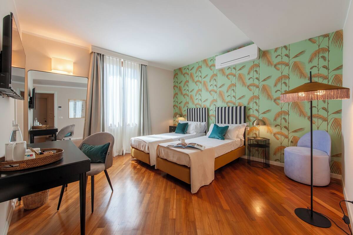 Une chambre d’hôtel avec deux lits, un bureau, une chaise, une lampe et un mur orné d’un motif de feuilles.