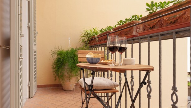 Un petit balcon avec une table dressée pour deux personnes, agrémentée d’un panier de pain, d’une tasse et de deux verres de vin rouge.