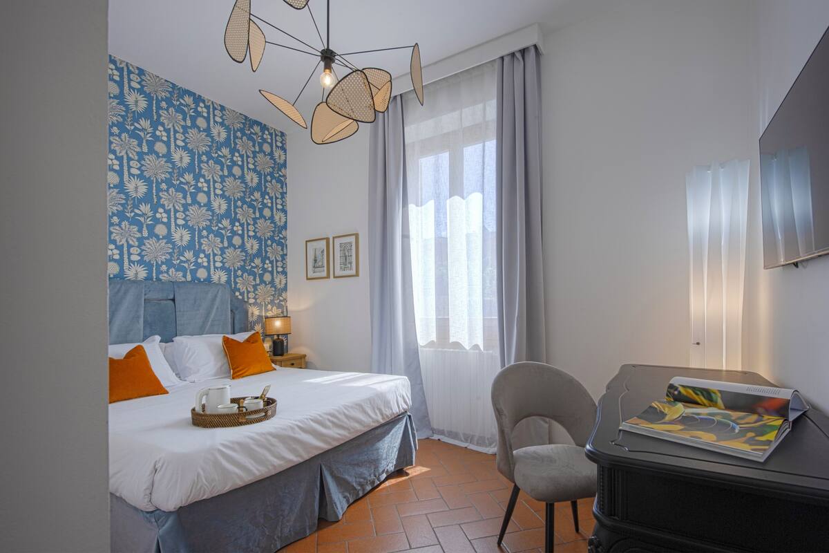 Une chambre d’hôtel comprenant un lit, un bureau, une chaise, une fenêtre avec des rideaux, un mur orné d’un motif floral et un luminaire au plafond.
