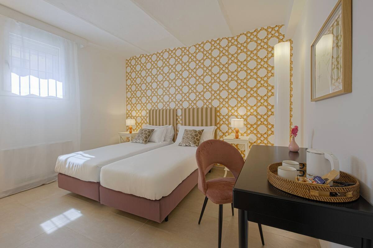 Un lit double avec une tête de lit rose, deux tables de chevet avec des lampes, une table d’appoint en osier et une chaise.