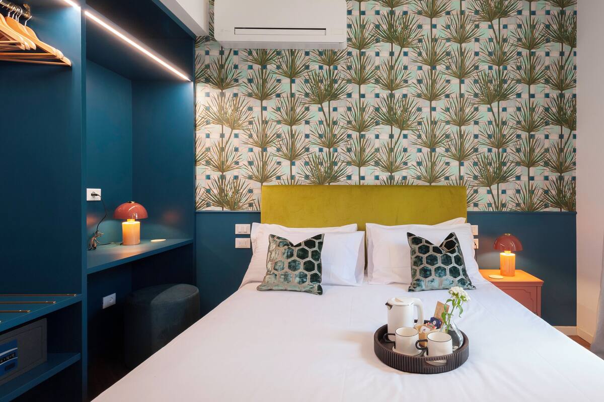 Une chambre d’hôtel avec un lit, une table de chevet, une lampe, un plateau avec des tasses et un mur orné d’un motif de feuilles.