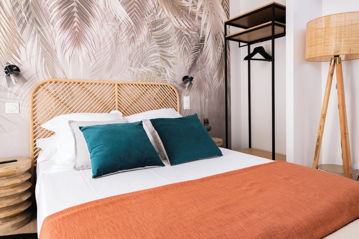 Ein ordentlich bezogenes Bett mit einem Holz-Kopfteil, zwei türkisfarbenen Kissen und einer orangefarbenen Tagesdecke.