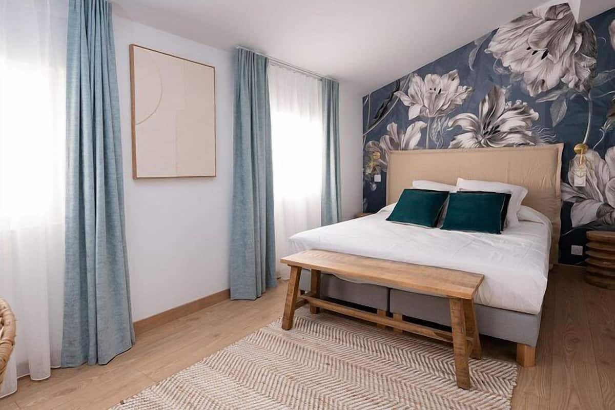 Ein Schlafzimmer mit einem Bett, einer Bank, einer blumengemusterten Wand und hellblauen Vorhängen.