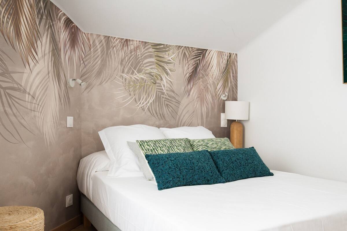 Ein Schlafzimmer mit einem Bett, weißer Bettwäsche, einem türkisfarbenen Zierkissen, einer Wandlampe und einem gewebten Korb.