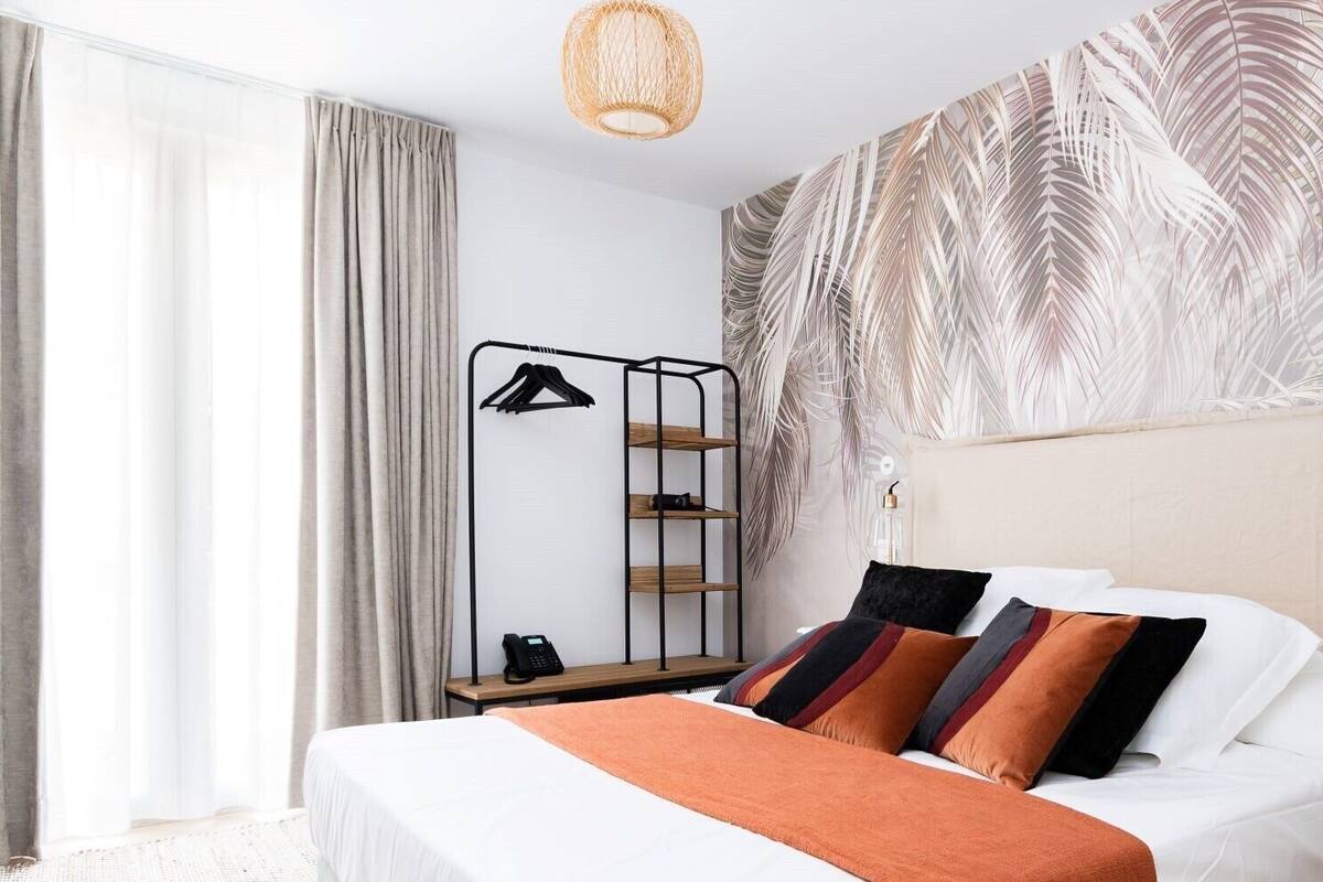 Ein modernes Hotelzimmer mit einem Bett, einem Nachttisch mit Telefon, einem wandmontierten Regal mit Kleiderhaken und einem großen Fenster mit Vorhängen.
