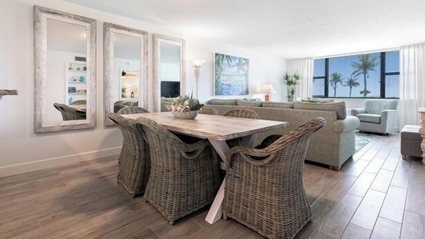 A modern living room with a wooden dining table, wicker chairs, a sofa, and a large window with a view of palm trees. - Direct Gulf Front Ground Floor Condo | Steps to the Gulf and Pet-Friendly (Sanibel)