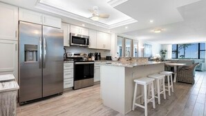 A modern kitchen with stainless steel appliances, a central island with bar stools, and a view of the outdoors through large windows. - Direct Gulf Front Ground Floor Condo | Steps to the Gulf and Pet-Friendly (Sanibel)