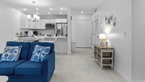 A modern living room with a blue sofa, a dining area, and a kitchen with white cabinets and appliances.