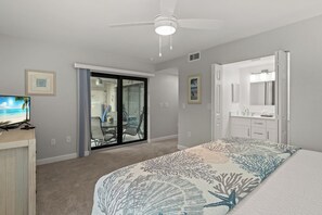 A bedroom with a bed, a ceiling fan, a sliding glass door leading to a balcony, and a television on a wooden stand.
