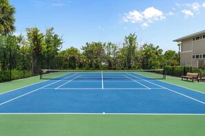 Una cancha de tenis al aire libre con red, rodeada de vegetación y un edificio.