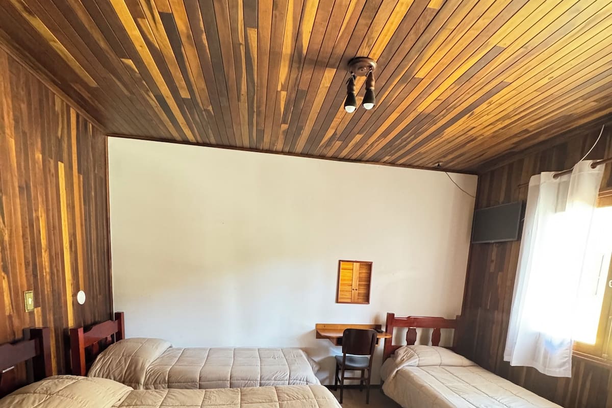 Una habitación con tres camas, techo de listones de madera, un escritorio pequeño y un televisor en la pared.