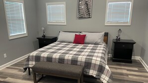 Ein Schlafzimmer mit einem Bett, einer Bank, zwei Nachttischen und drei Fenstern mit Jalousien.