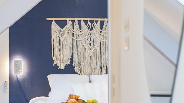 Un dormitorio con una cama, un colgador de macramé en la pared y una mesita con frutas y pasteles.