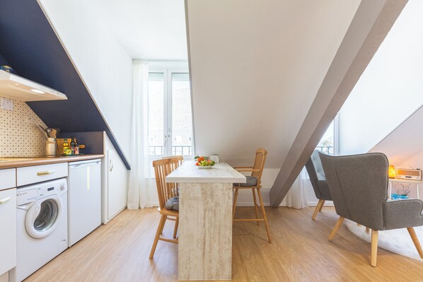 A kitchen with a central island, wooden chairs, a washing machine, and a window with curtains. - Le Perchoir, with parking, across from the thermal baths (Bagnères-de-Luchon)