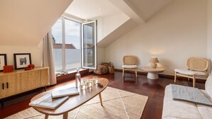 A spacious loft apartment with a high ceiling, a large skylight, a wooden dining table, a white sofa, and a rattan chair.