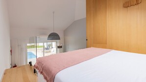 Een slaapkamer met een groot bed, een hanglamp, een houten deur en uitzicht op een buitenzwembad.