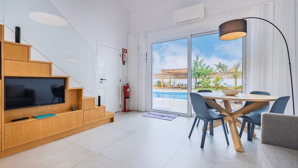 Een moderne woonkamer met een flatscreen televisie, houten meubels, een eethoek met een ronde tafel en stoelen, en een grote schuifglazen deur die leidt naar een buitenzwembad.