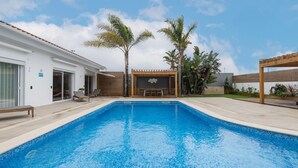 Ein moderner Außenpool mit einer Holzpergola und Liegestühlen, umgeben von tropischen Pflanzen und einem weißen Gebäude mit einer Schiebeglastür.