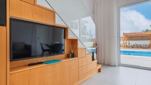 Ein modernes Wohnzimmer mit großem Flachbildfernseher, Holzschränken, einem kleinen Tisch mit einer Vase und Blick auf einen Außenpool.