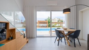 Ein modernes Wohnzimmer mit Glastür zum Poolbereich, einem Esstisch mit Stühlen und einem Fernseher auf einer Holzkommode.