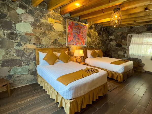 A room with two beds, stone walls, wooden beams, a hanging light fixture, and a window with curtains.