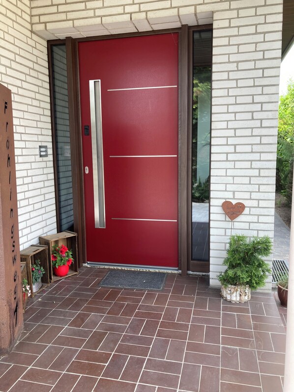 Una puerta roja con un picaporte vertical, un pequeño banco de madera y una maceta roja con flores, además de una decoración con forma de corazón en la pared.