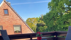 A wooden deck with two black chairs and a small table, a red flower pot, and a brick building in the background.