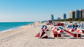 Ein Strand mit Reihen aus roten und weißen Sonnenschirmen und Liegestühlen, einem Rettungsschwimmturm und hohen Gebäuden im Hintergrund.