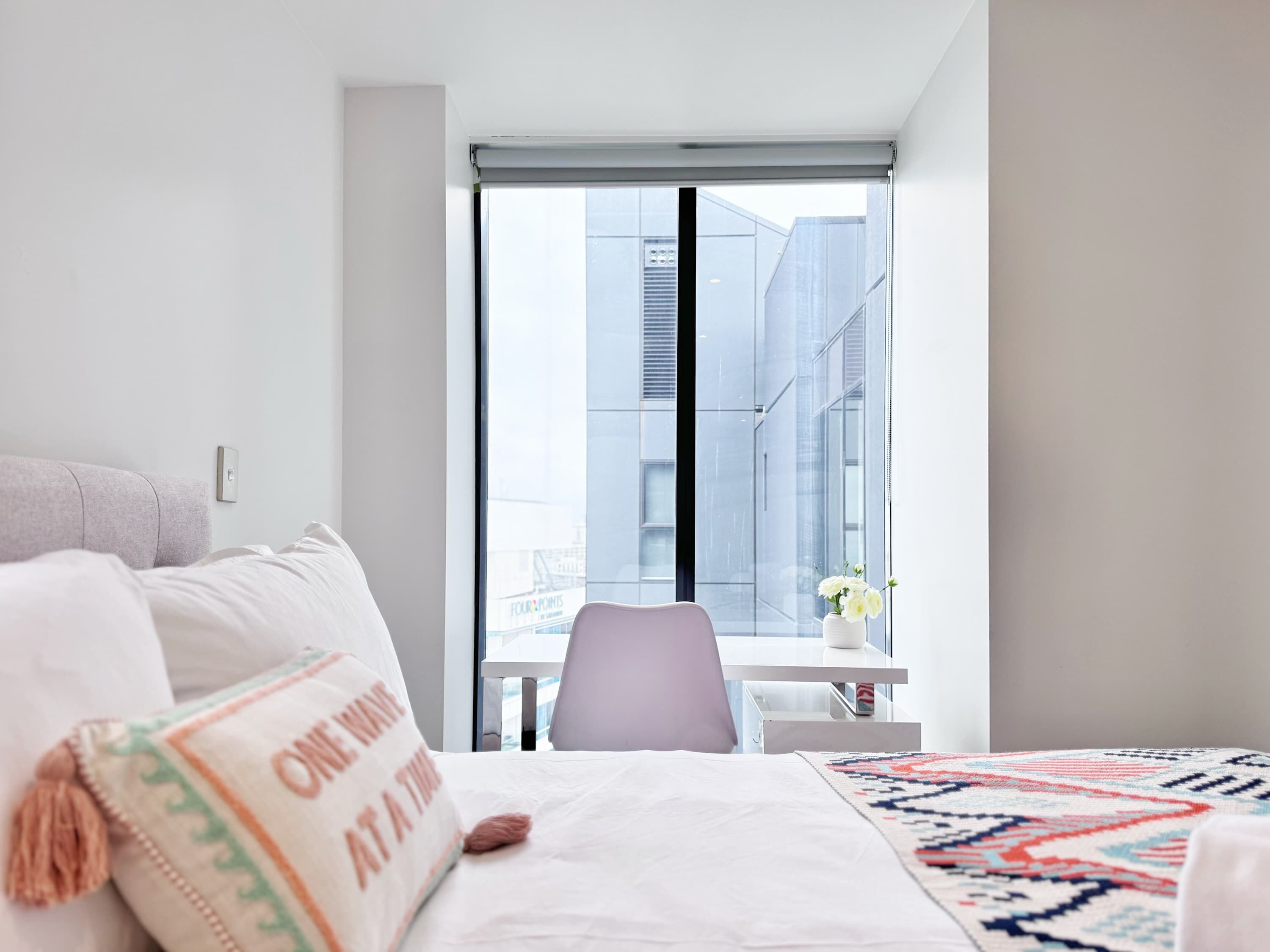 Una habitación de hotel moderna con un ventanal grande, una cama con ropa blanca, un escritorio pequeño y una silla.
