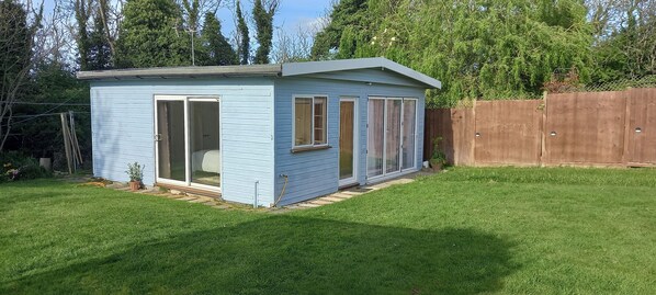 A small, light blue shed with a sliding glass door and windows, situated in a grassy backyard with a wooden fence in the background.