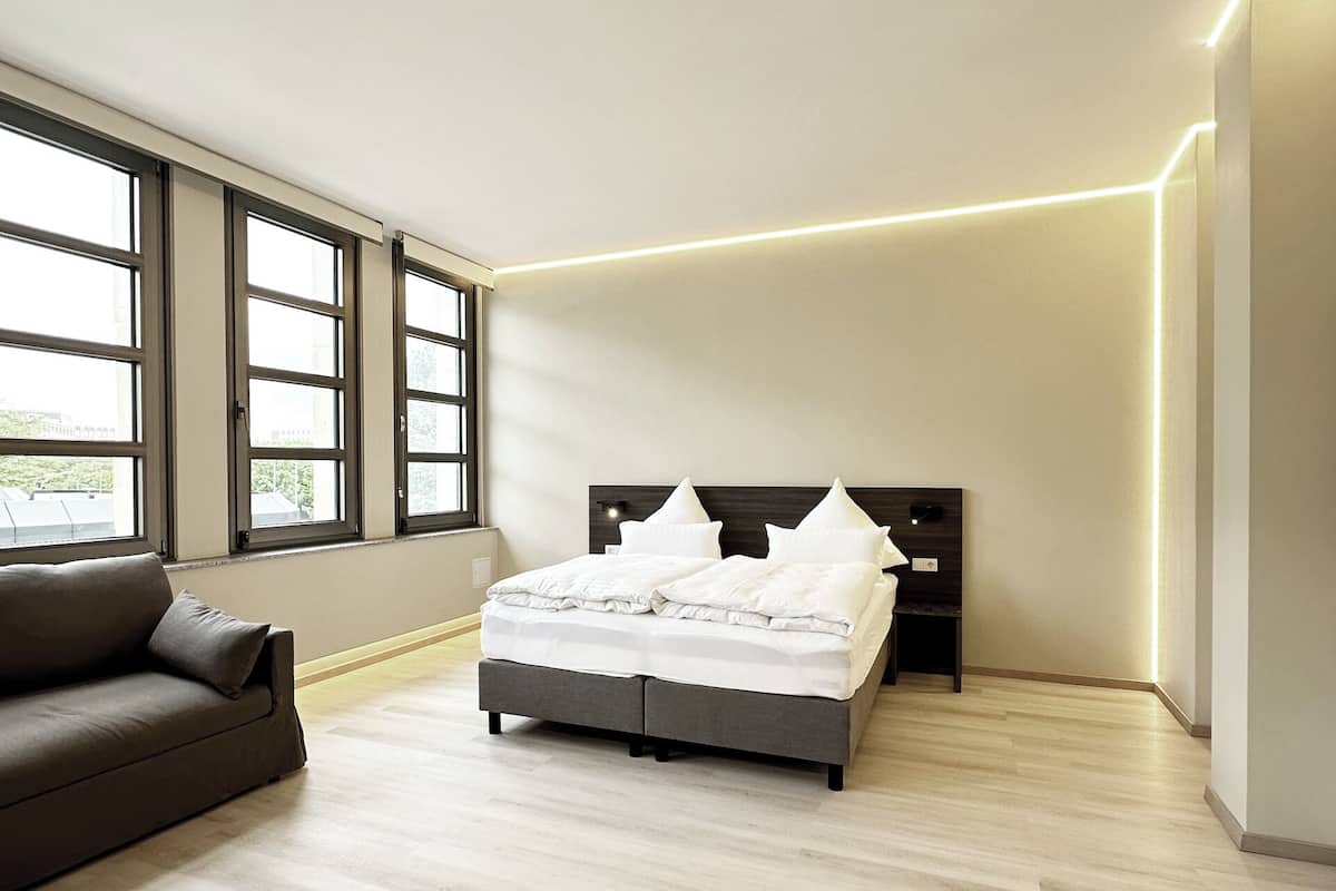 A modern bedroom with a bed, a sofa, and large windows.