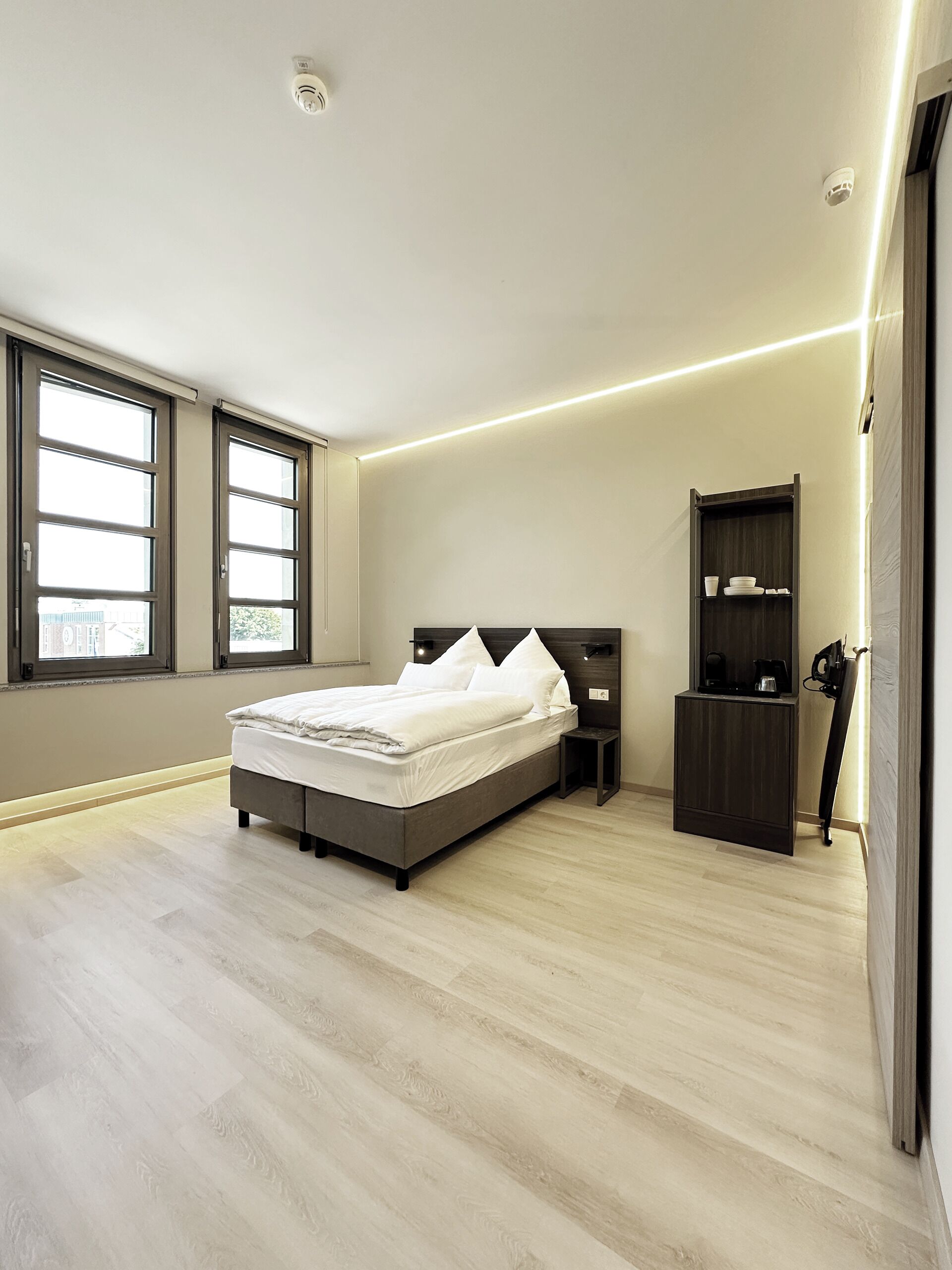 A modern bedroom with a large bed, built-in nightstands, a dark wooden wardrobe, and a large window providing natural light.